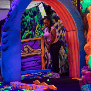 Woman putting on an indoor mini golf course under an archway that looks like a shark mouth.