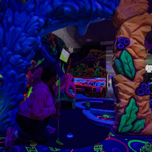 Woman kneeling down next to indoor mini golf tee to line up her shot.