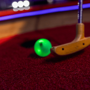 Green glowing golf ball being putted into mini golf cup by putter on red carpet.