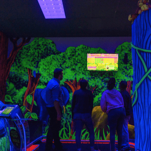 4 people with their backs to the camera looking up at a Putt Mania scoreboard on an indoor mini golf course.