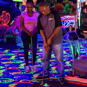 A man putting off a tee on an indoor mini golf Putt Mania course with a woman watching him.