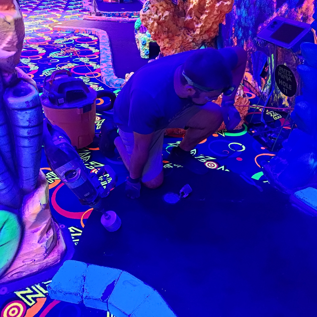 Man bent down wearing PPE next to a hole on a mini golf course.