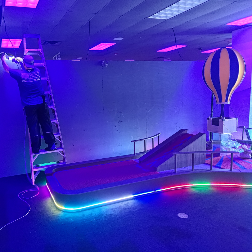 Man standing on a ladder in a construction setting on an indoor mini golf course.