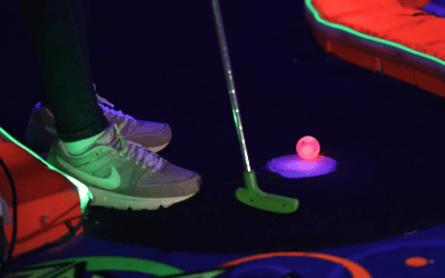 Person's feet in tennis shoes standing next to a mini golf tee with red illuminated Putt Mania golf ball