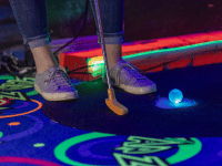 Person's feet in tennis shoes standing next to a mini golf tee with blue illuminated Putt Mania golf ball