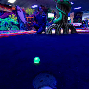 Putt Mania illuminated green golf ball next to a cup on an indoor mini golf course with a woman knealing down and watching in the background.