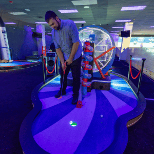 A man putting a golf ball into a cup on a ferris wheel theme hole on an indoor mini golf course.