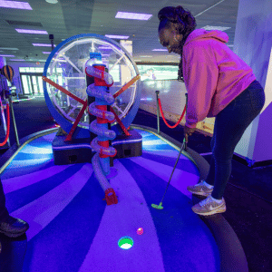 A woman putting a golf ball into a cup on a ferris wheel theme hole on an indoor mini golf course.