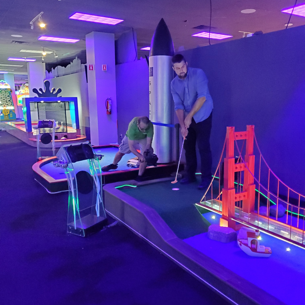 Man putting on a mini golf course with another man bent down taking a photo of him.