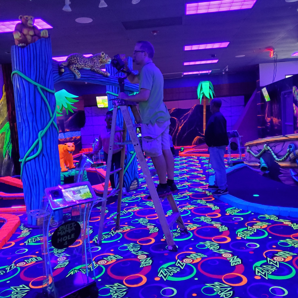 Man in a green shirt and shorts standing on a ladder at a glow in the dark mini golf course holding a camera.