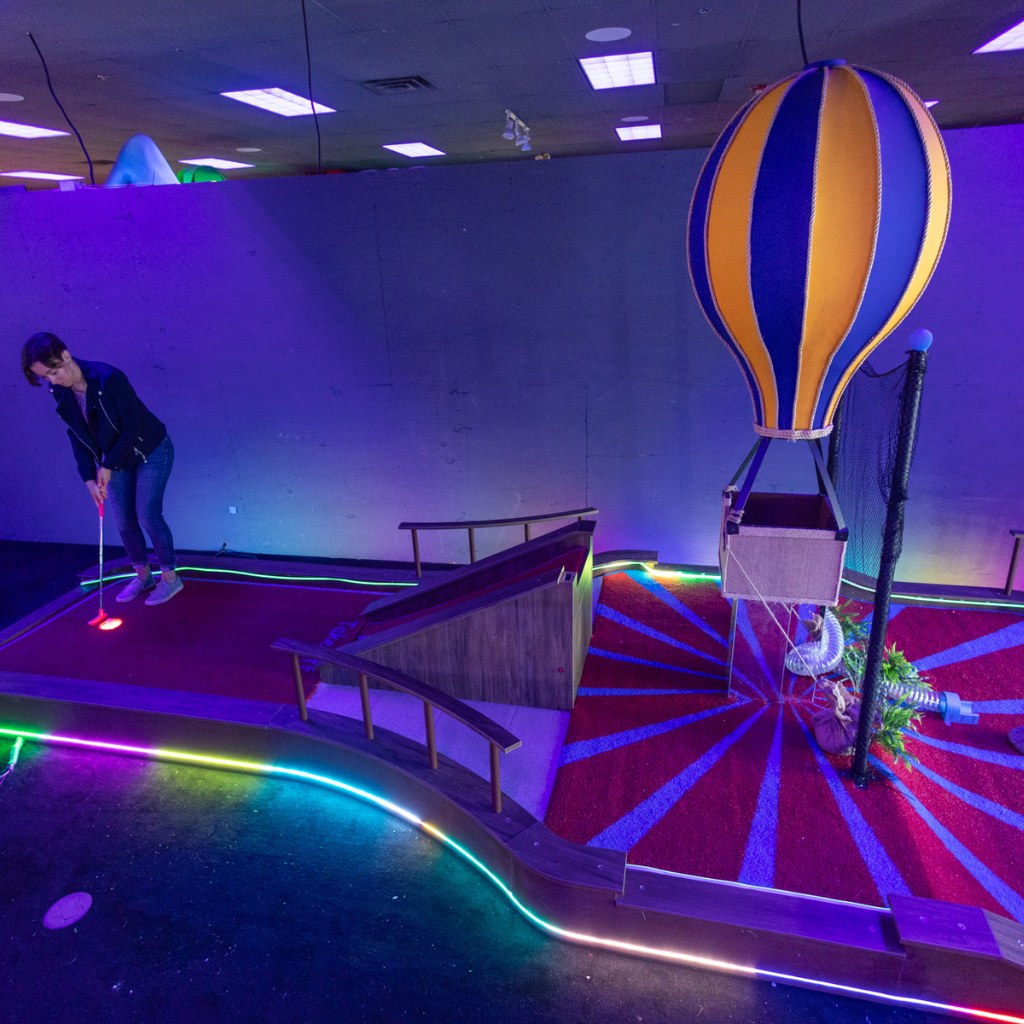 Person putting on a lit up indoor mini golf course with a hot air balloon prop.