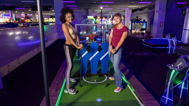 Two women standing on a soccer theme mini golf hole, smiling at the camera.