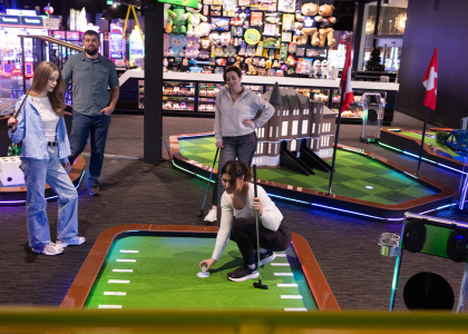 Three people standing around an indoor mini golf course with another person places a ball down on the tee.