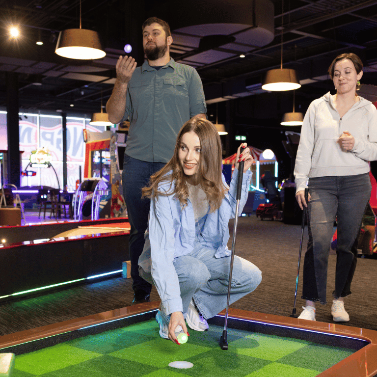 A woman putting a golf ball on the tee at an indoor mini golf course with a man and a wowman spectating behind her.